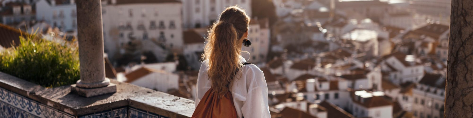 Blonde woman standing on the balcony and looking at coast view of the southern european city with sea during the sunset, wearing hat, cork bag, safari shorts and white shirt